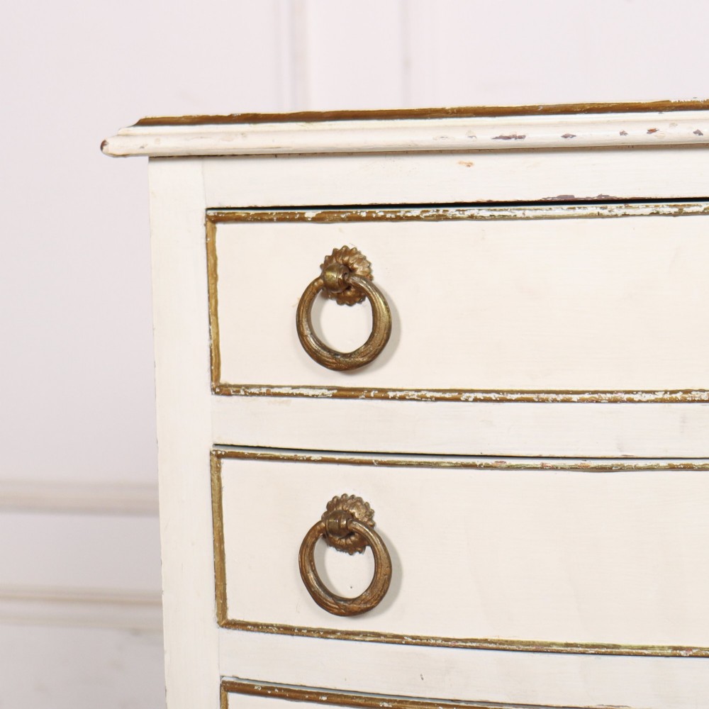 A pair of off-white painted French bedside chests featuring three curved drawers, gilded moulding details, brass ring handles, and tall square tapered legs. View IV