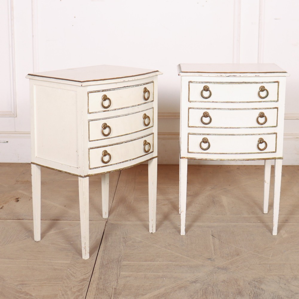 A pair of off-white painted French bedside chests featuring three curved drawers, gilded moulding details, brass ring handles, and tall square tapered legs. View V