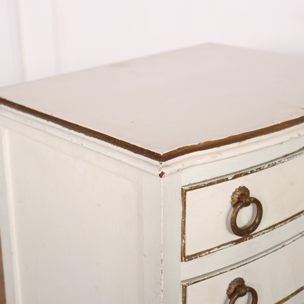A pair of off-white painted French bedside chests featuring three curved drawers, gilded moulding details, brass ring handles, and tall square tapered legs. View VI