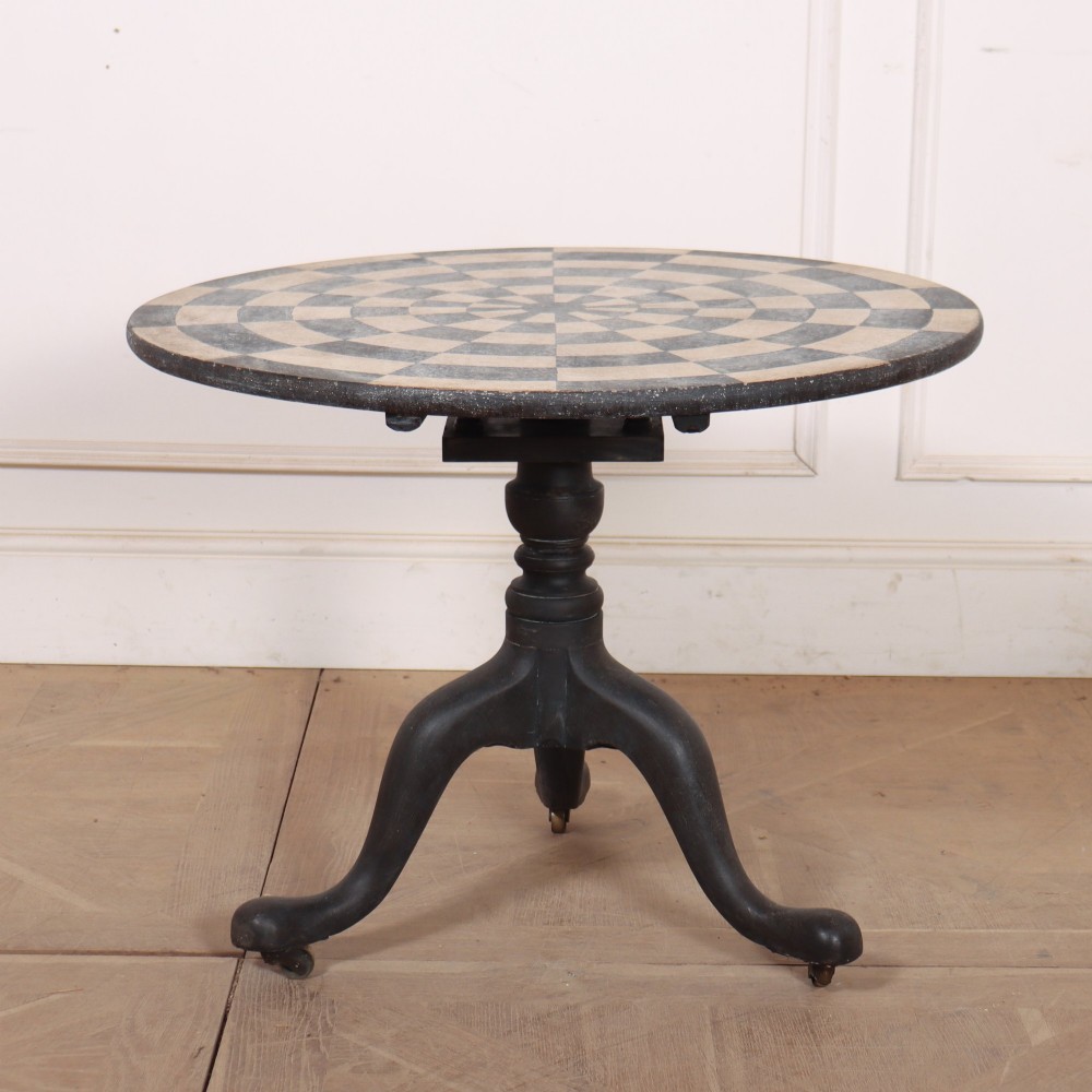 A circular tilt-top tripod table featuring a painted concentric checkerboard top in black and cream, supported by a dark three-legged base on castors. View VII