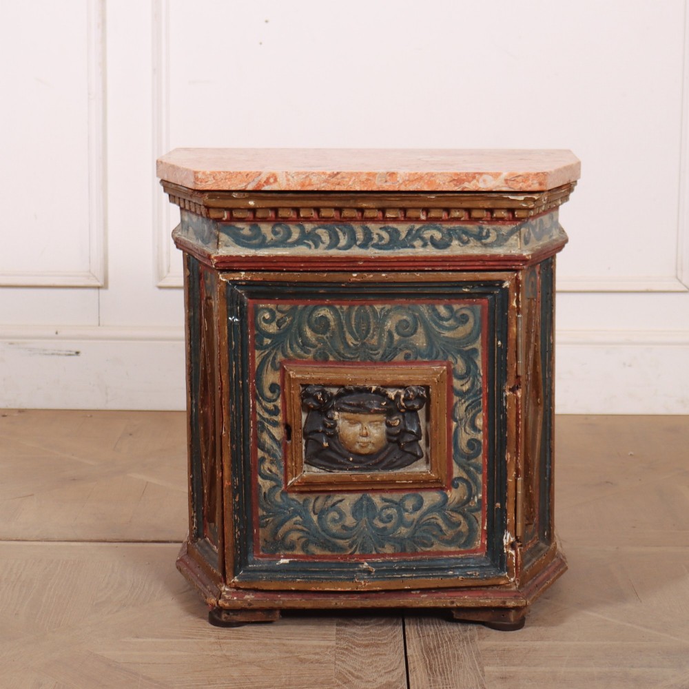An antique Italian polychrome painted cabinet with a pink marble top, canted corners, and a central carved face mask panel.