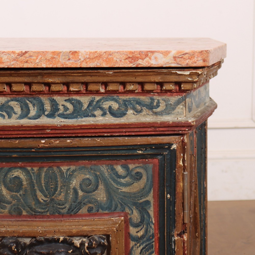 An antique Italian polychrome painted cabinet with a pink marble top, canted corners, and a central carved face mask panel. View IV