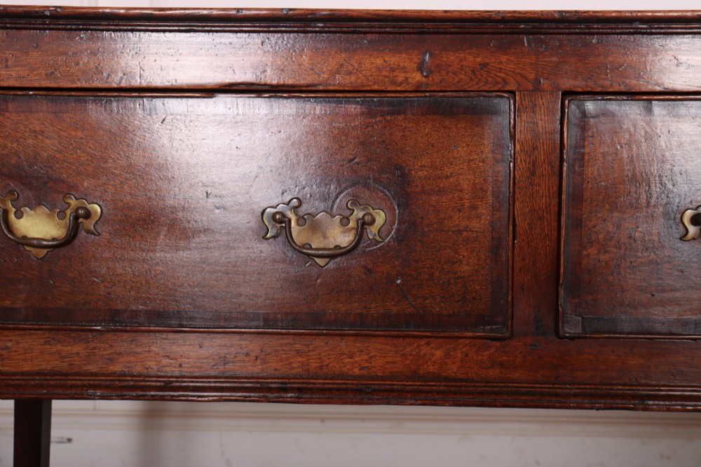 An eighteenth-century English oak dresser base featuring three drawers with brass swan-neck handles, raised on four slender straight legs. View V
