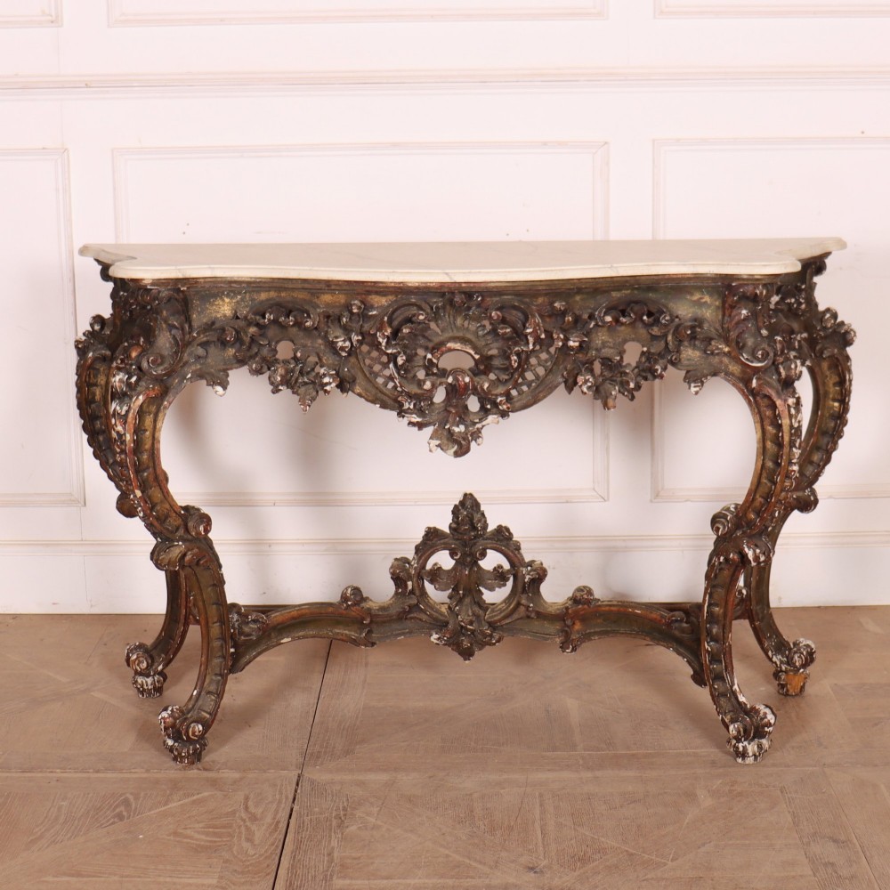 An antique French Rococo style console table featuring a cream marble top, elaborate rocaille carved giltwood apron, cabriole legs, and a decorative central stretcher.