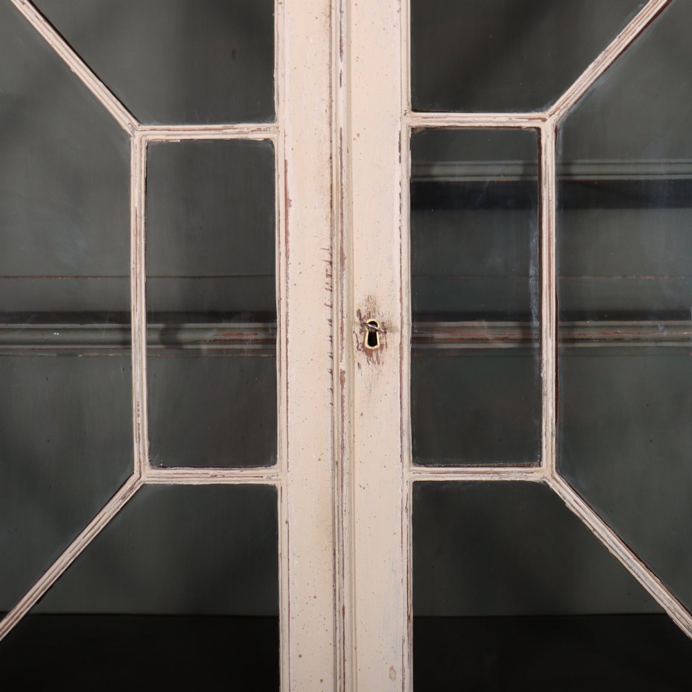A cream painted English glazed bookcase featuring upper doors with geometric astragal glazing bars above a solid two-door cupboard base. View IV
