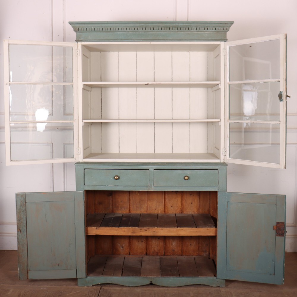 A tall two-part painted muted blue wood cabinet featuring an upper section with two glazed glass doors over a lower cupboard base with two drawers and panelled doors. View X