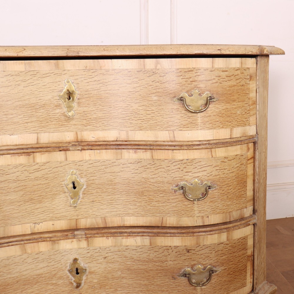 An Austrian bleached oak commode featuring a serpentine front, three drawers with crossbanded borders, brass escutcheons, handles, and turned feet. View V
