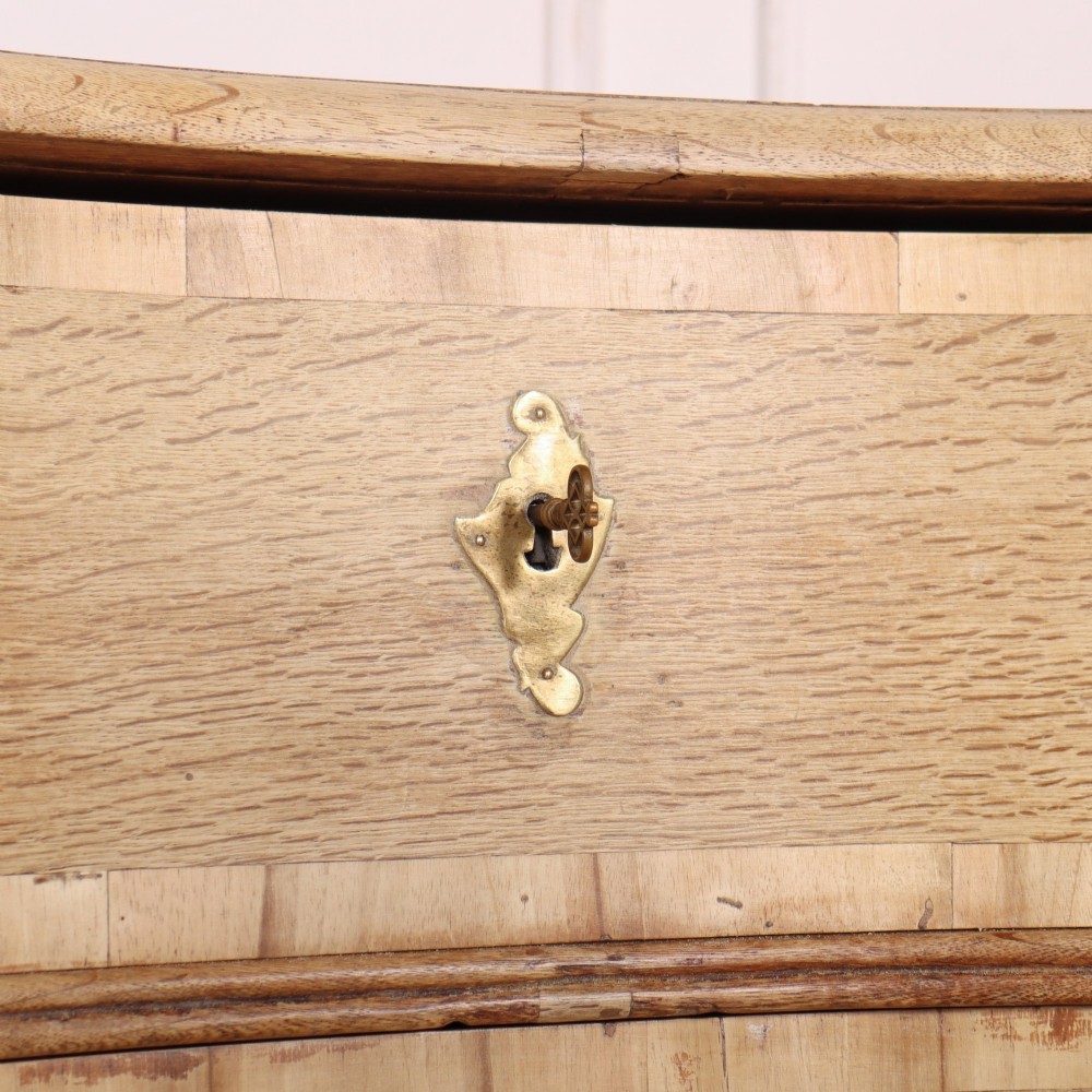 An Austrian bleached oak commode featuring a serpentine front, three drawers with crossbanded borders, brass escutcheons, handles, and turned feet. View VI