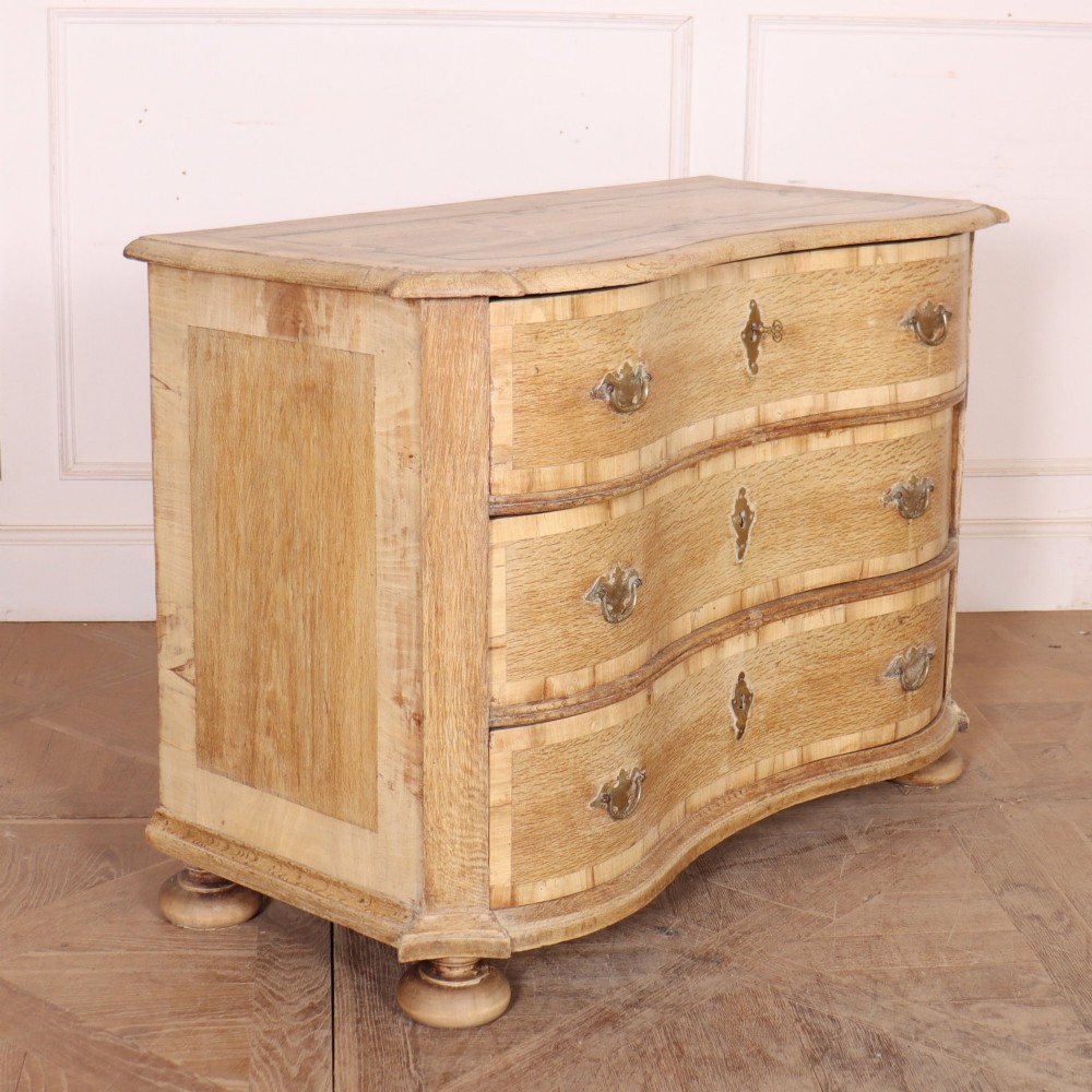 An Austrian bleached oak commode featuring a serpentine front, three drawers with crossbanded borders, brass escutcheons, handles, and turned feet. View VII
