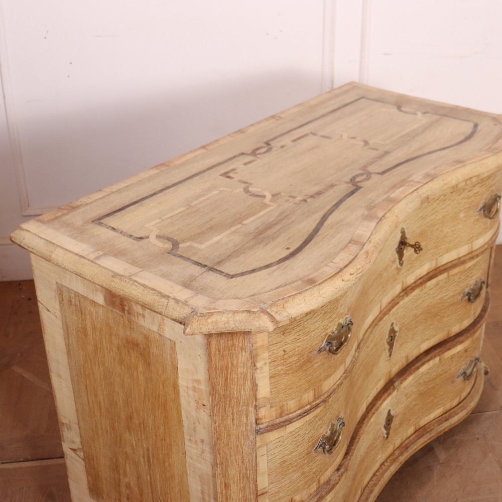 An Austrian bleached oak commode featuring a serpentine front, three drawers with crossbanded borders, brass escutcheons, handles, and turned feet. View X