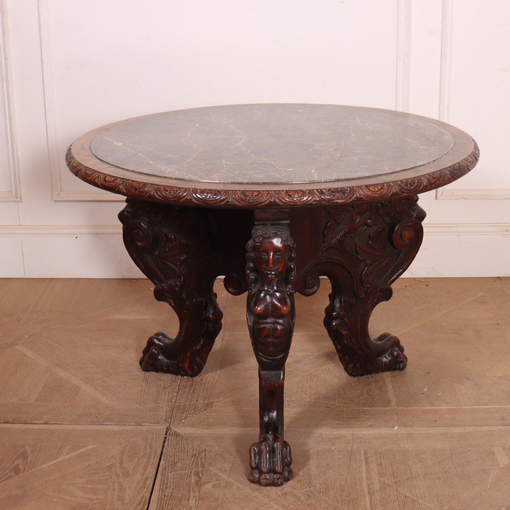 A circular dark wood lamp table featuring an inset dark marble top, intricately carved floral borders, and carved caryatid legs resting on paw feet.