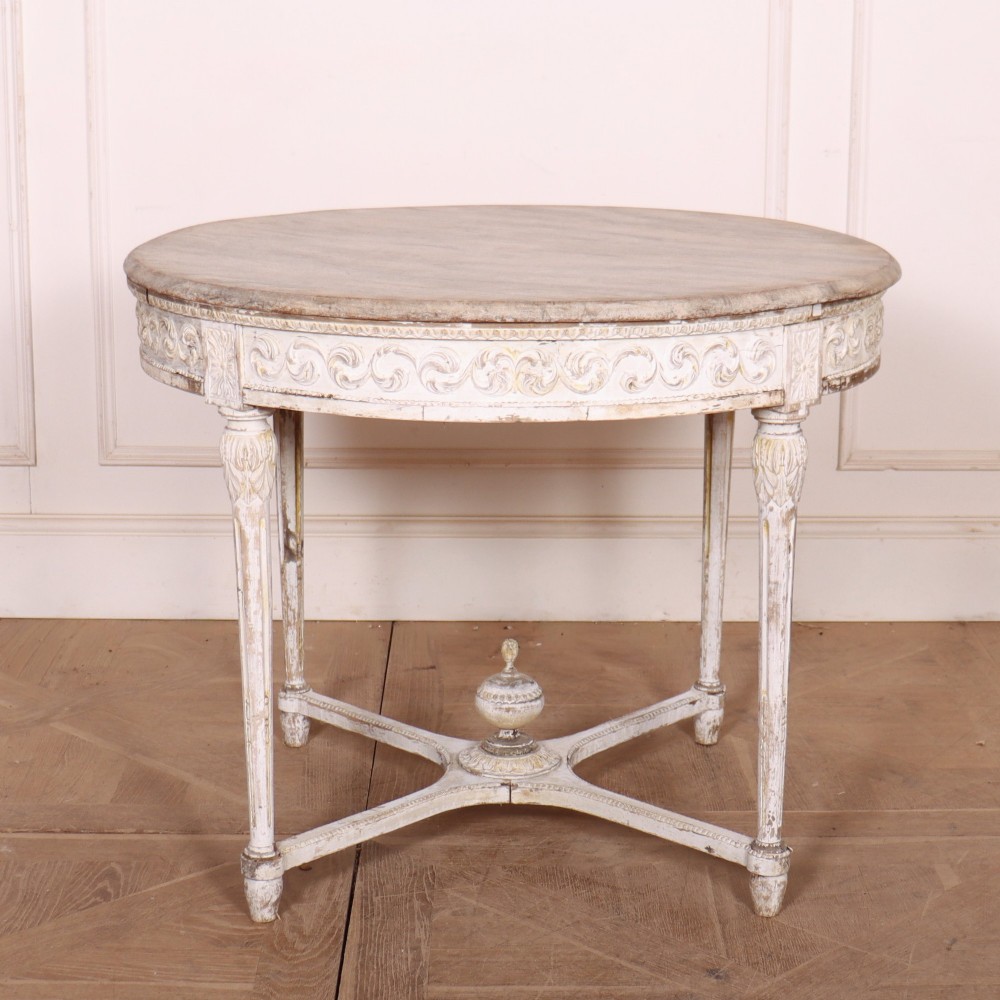 A Swedish Gustavian style circular side table featuring a distressed off-white painted finish, carved wave pattern apron, fluted legs, and an X-shaped stretcher with a central urn finial.