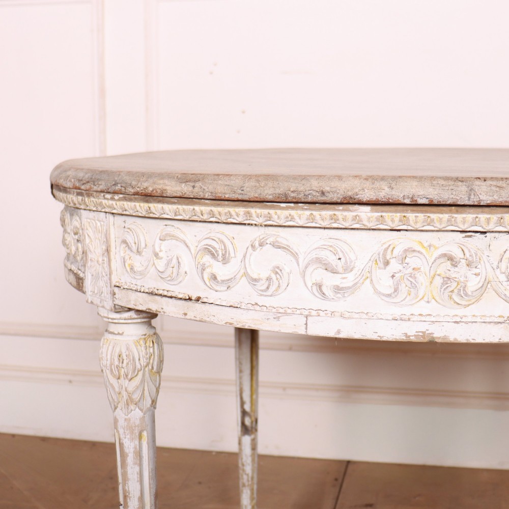 A Swedish Gustavian style circular side table featuring a distressed off-white painted finish, carved wave pattern apron, fluted legs, and an X-shaped stretcher with a central urn finial. View II