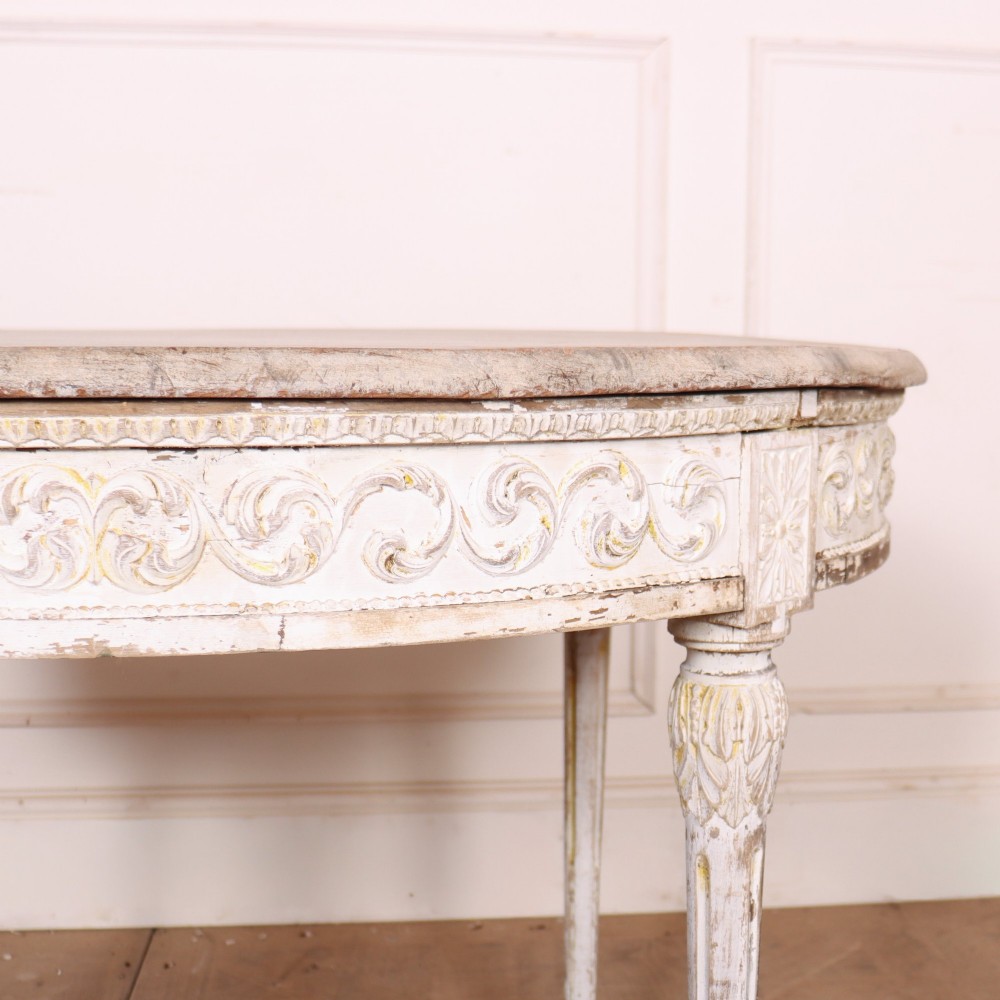 A Swedish Gustavian style circular side table featuring a distressed off-white painted finish, carved wave pattern apron, fluted legs, and an X-shaped stretcher with a central urn finial. View III
