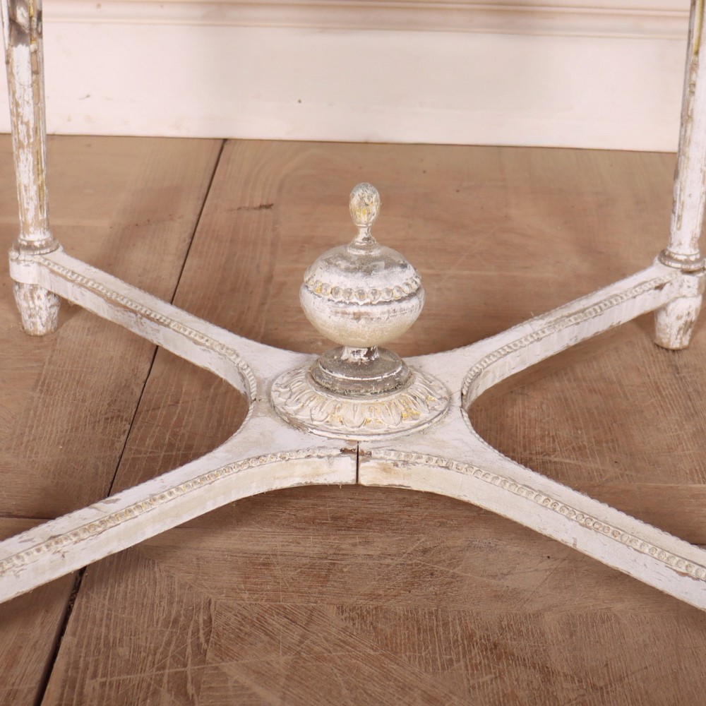 A Swedish Gustavian style circular side table featuring a distressed off-white painted finish, carved wave pattern apron, fluted legs, and an X-shaped stretcher with a central urn finial. View IV