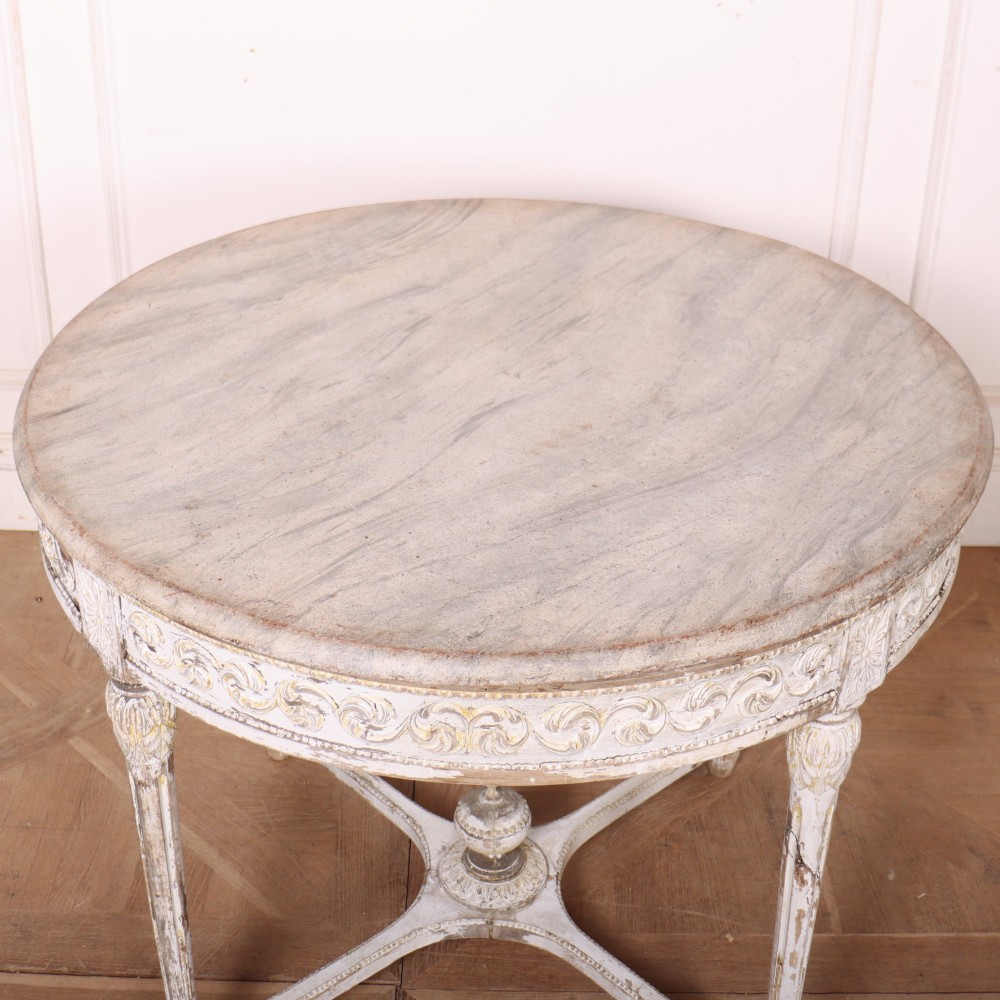 A Swedish Gustavian style circular side table featuring a distressed off-white painted finish, carved wave pattern apron, fluted legs, and an X-shaped stretcher with a central urn finial. View V
