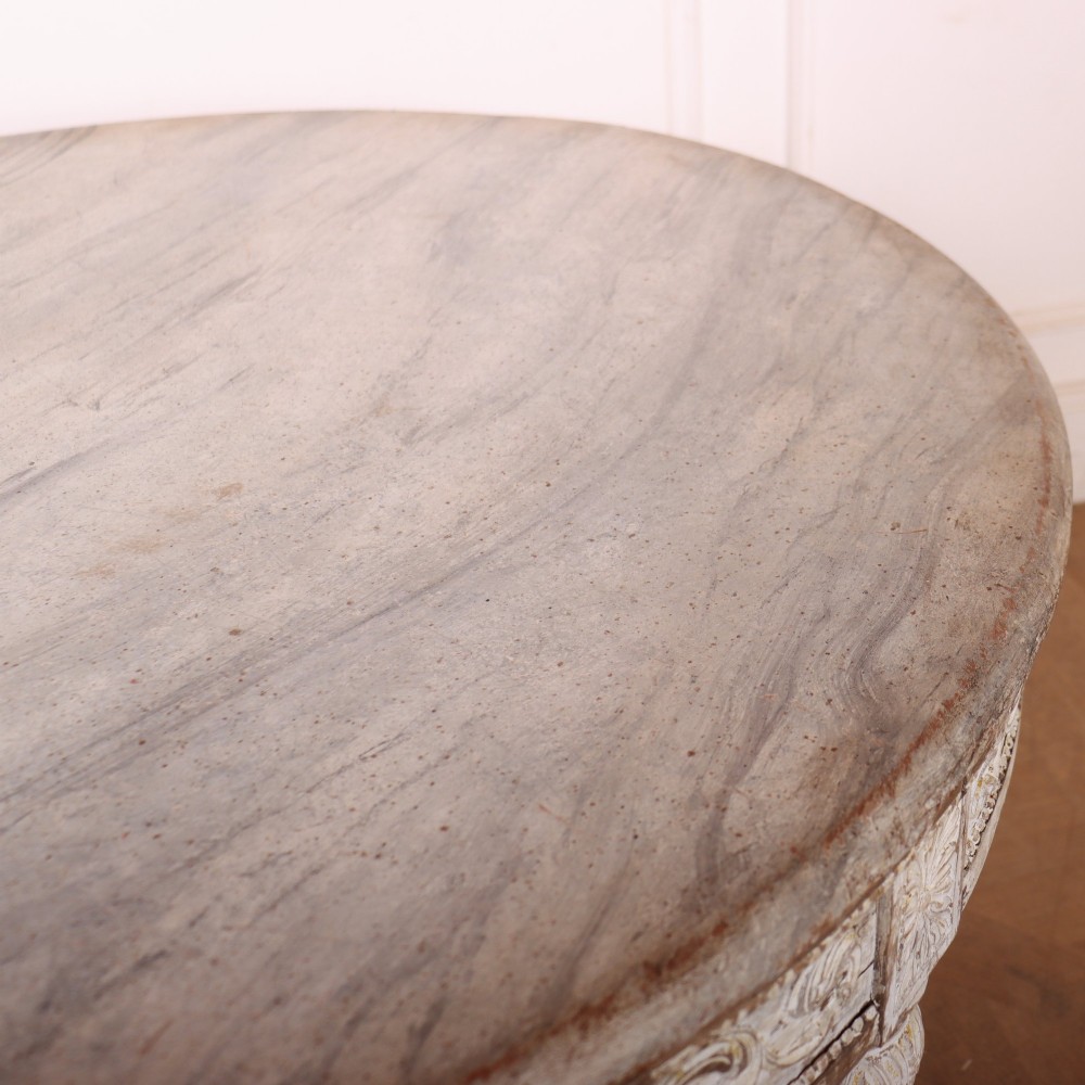 A Swedish Gustavian style circular side table featuring a distressed off-white painted finish, carved wave pattern apron, fluted legs, and an X-shaped stretcher with a central urn finial. View VII