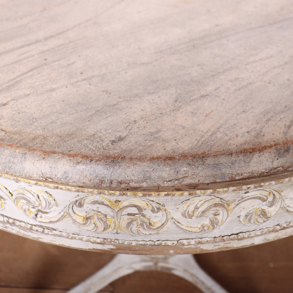 A Swedish Gustavian style circular side table featuring a distressed off-white painted finish, carved wave pattern apron, fluted legs, and an X-shaped stretcher with a central urn finial. View VIII