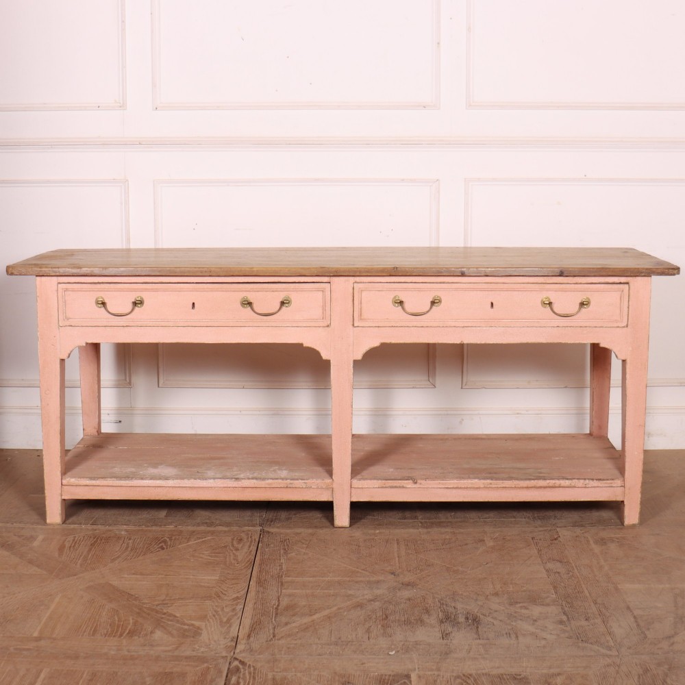 A Scottish farmhouse potboard dresser base with a painted pink frame, wide drawers fitted with brass handles, and an open lower wooden shelf.