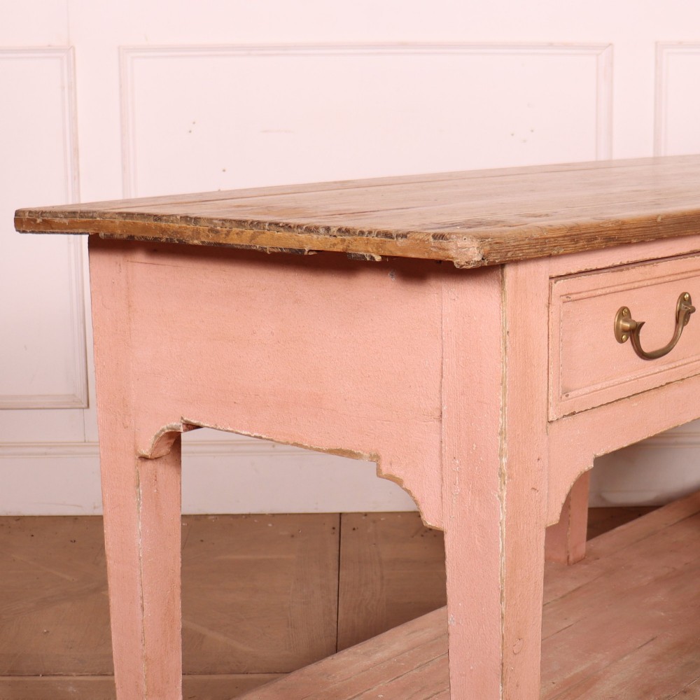 A Scottish farmhouse potboard dresser base with a painted pink frame, wide drawers fitted with brass handles, and an open lower wooden shelf. View X
