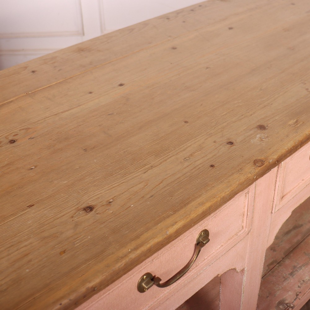 A Scottish farmhouse potboard dresser base with a painted pink frame, wide drawers fitted with brass handles, and an open lower wooden shelf. View XII
