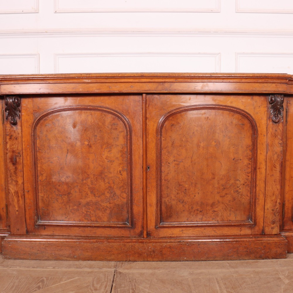 An antique English burr walnut dresser base featuring six arched cupboard doors with hand-carved decorative corbels along the top moulding. View IX