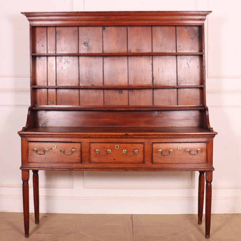 An antique English pine kitchen dresser featuring an open tiered plate rack above three drawers with brass handles, resting on slender turned legs.