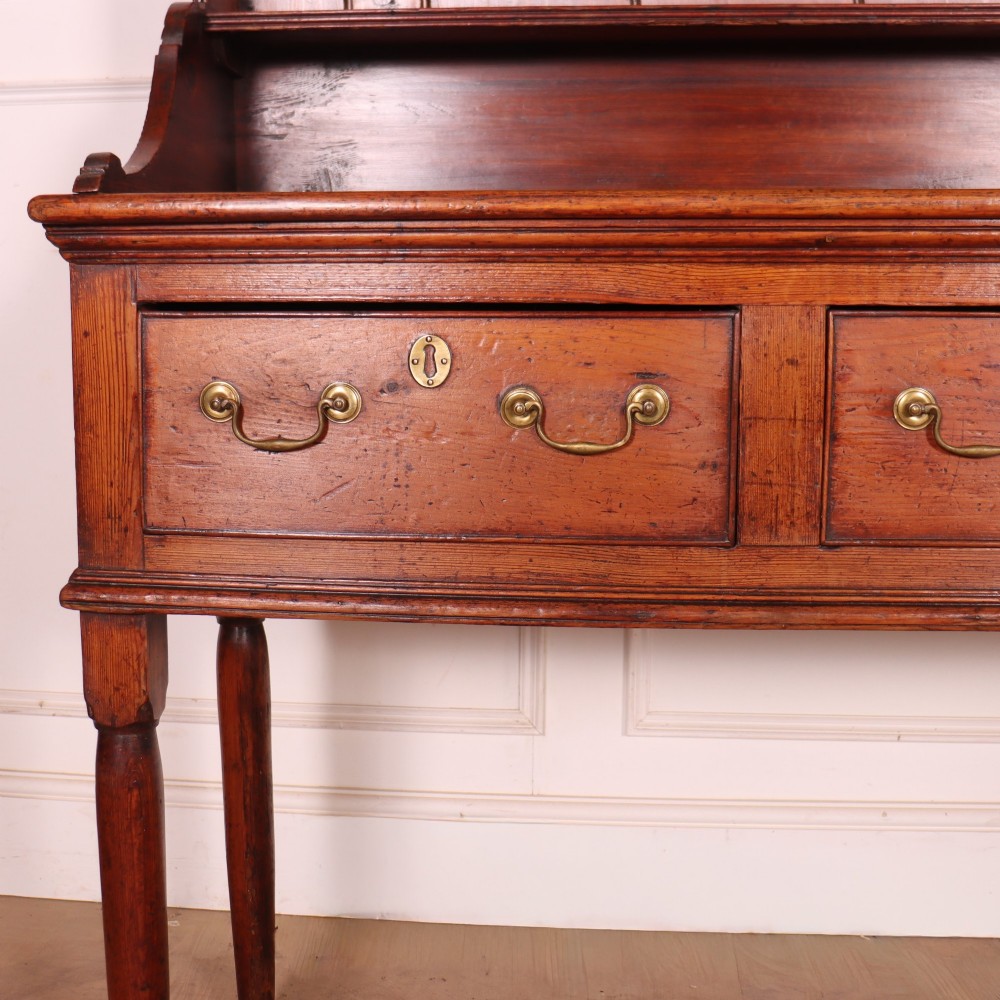 An antique English pine kitchen dresser featuring an open tiered plate rack above three drawers with brass handles, resting on slender turned legs. View II