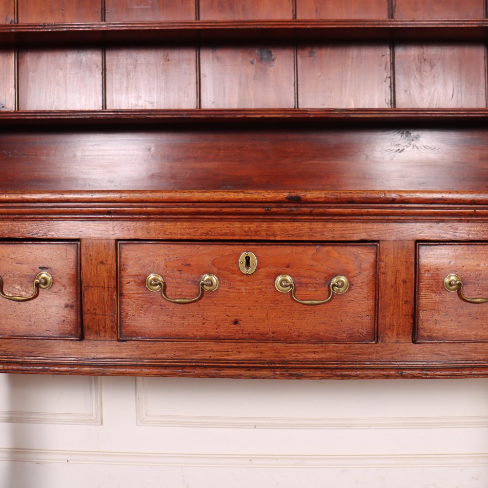 An antique English pine kitchen dresser featuring an open tiered plate rack above three drawers with brass handles, resting on slender turned legs. View III