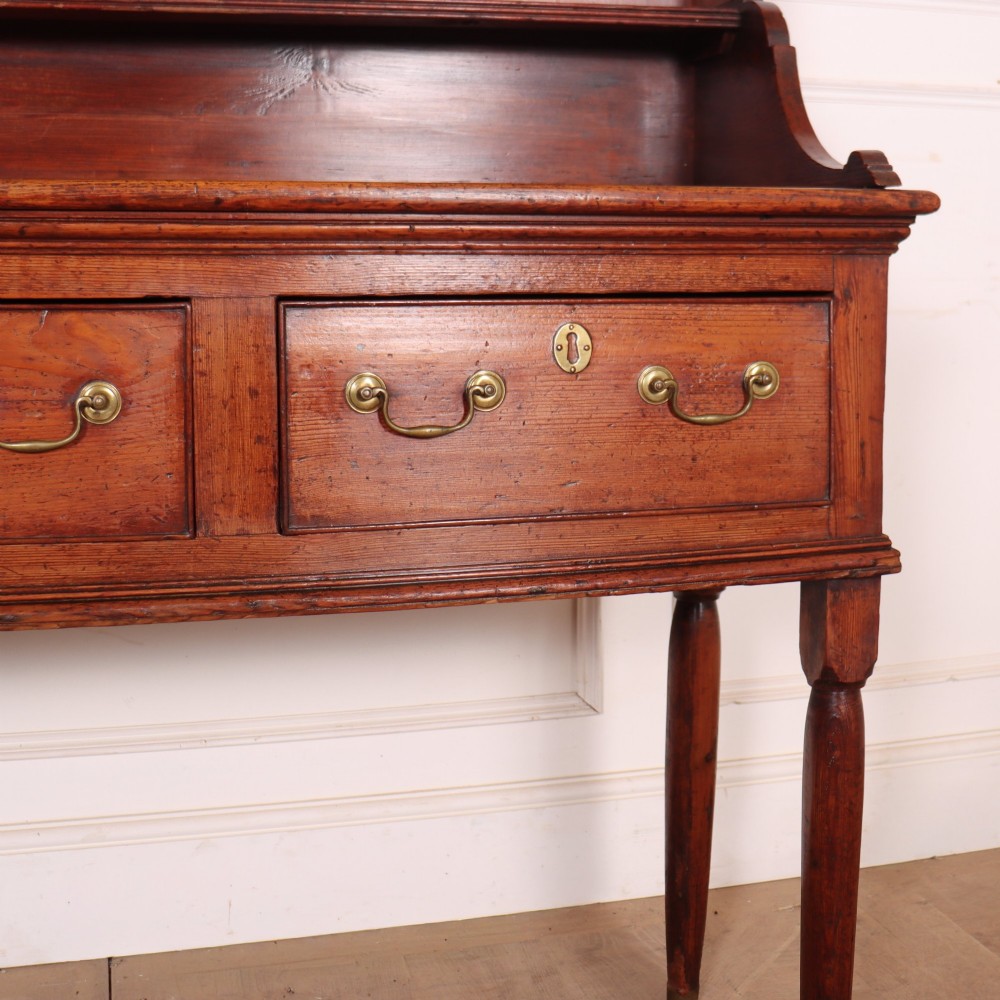 An antique English pine kitchen dresser featuring an open tiered plate rack above three drawers with brass handles, resting on slender turned legs. View IV