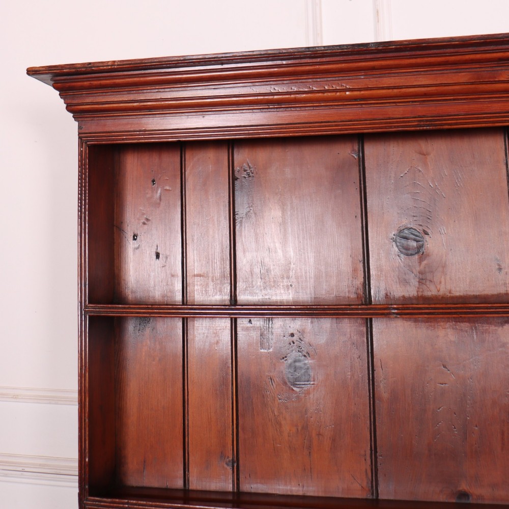 An antique English pine kitchen dresser featuring an open tiered plate rack above three drawers with brass handles, resting on slender turned legs. View VI