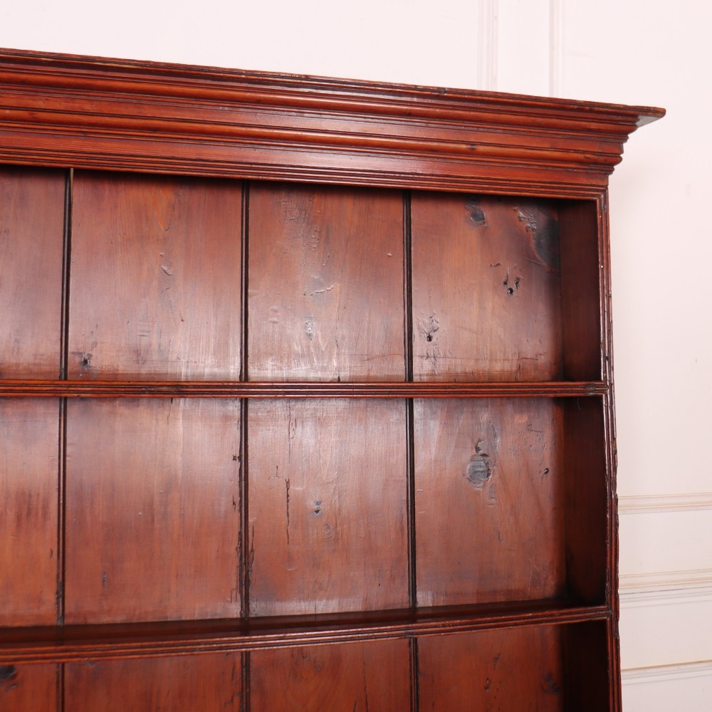 An antique English pine kitchen dresser featuring an open tiered plate rack above three drawers with brass handles, resting on slender turned legs. View VII