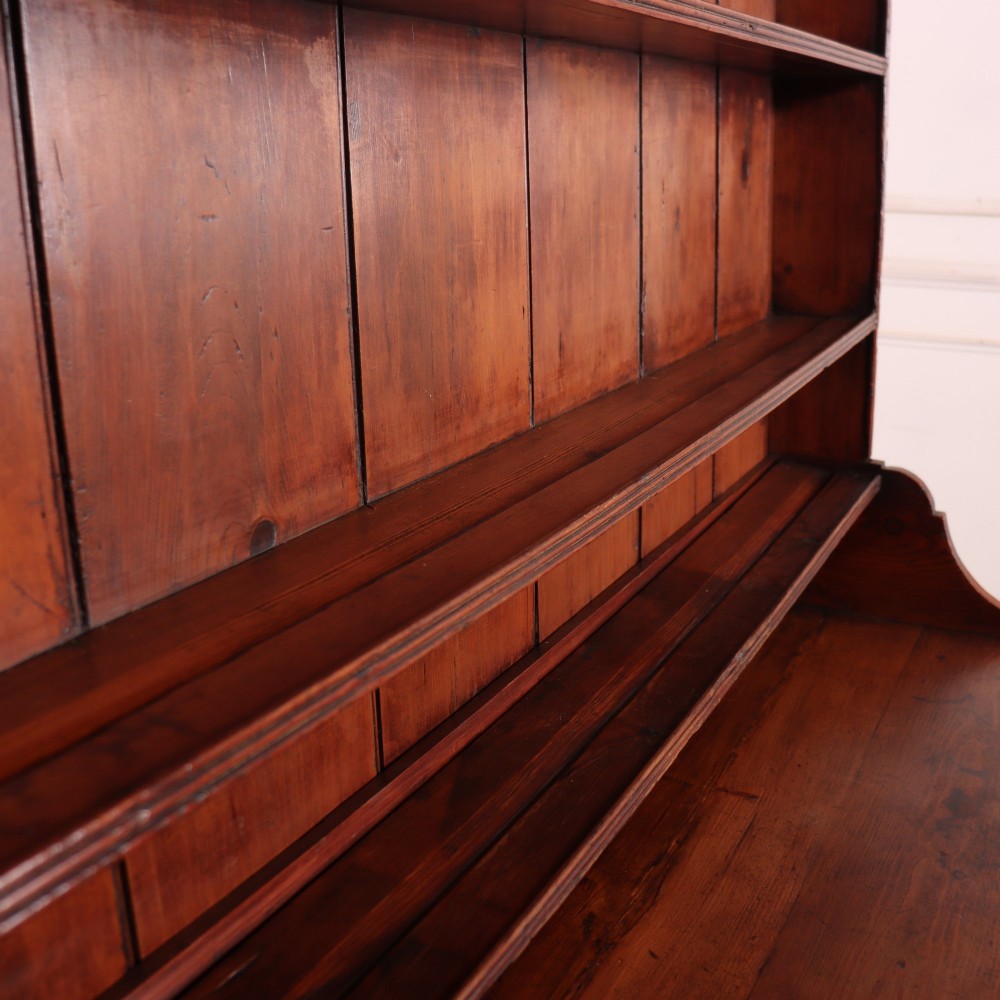 An antique English pine kitchen dresser featuring an open tiered plate rack above three drawers with brass handles, resting on slender turned legs. View X