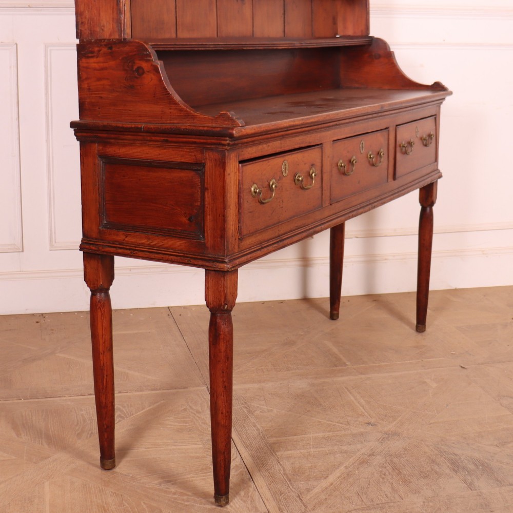 An antique English pine kitchen dresser featuring an open tiered plate rack above three drawers with brass handles, resting on slender turned legs. View XI