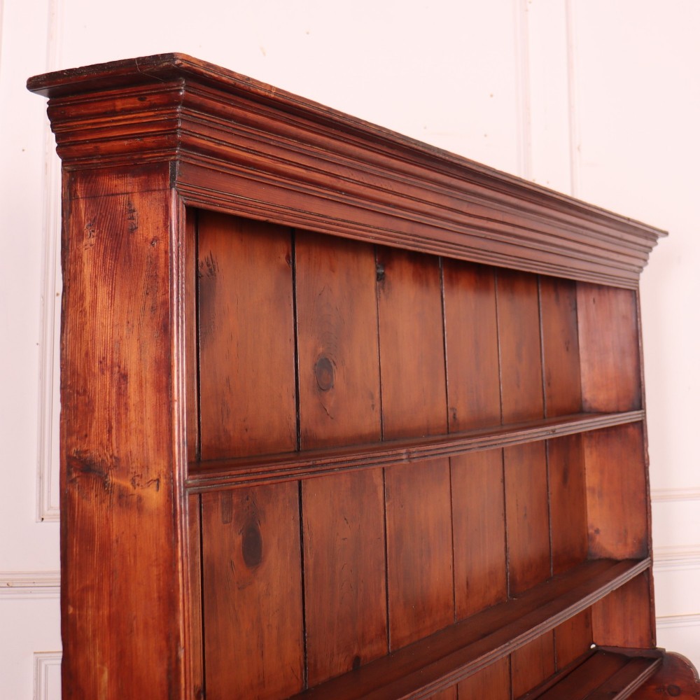 An antique English pine kitchen dresser featuring an open tiered plate rack above three drawers with brass handles, resting on slender turned legs. View XII