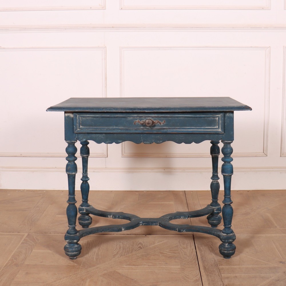 A rectangular blue painted wooden lamp table featuring turned legs, a single drawer with a metal pull, a scalloped apron, and a curved X-shaped stretcher.