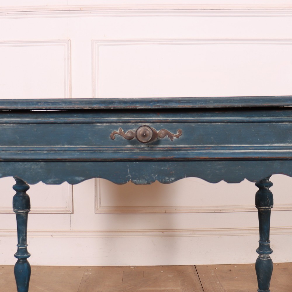A rectangular blue painted wooden lamp table featuring turned legs, a single drawer with a metal pull, a scalloped apron, and a curved X-shaped stretcher. View VI