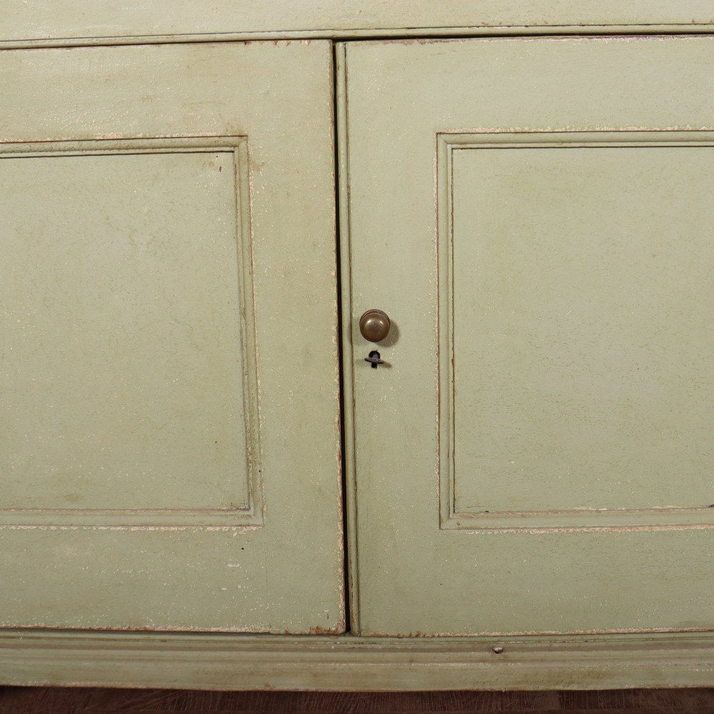 A large sage green painted dresser featuring an upper glazed display cabinet with sliding doors, lower drawers with brass drop handles, and panelled cupboard doors. View VII
