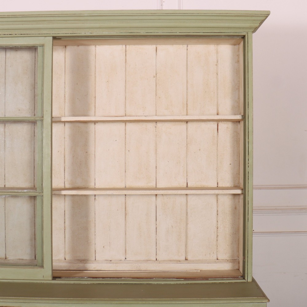 A large sage green painted dresser featuring an upper glazed display cabinet with sliding doors, lower drawers with brass drop handles, and panelled cupboard doors. View X