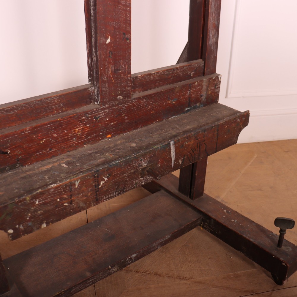 A floor-standing antique dark oak studio easel featuring an adjustable vertical central mast and a wide H-frame base with castors. View V