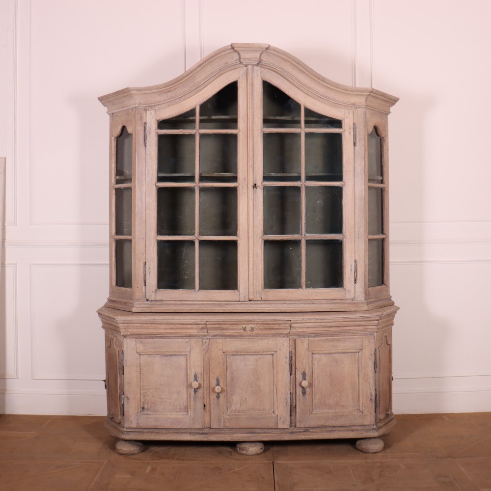 A large Dutch bleached oak display cabinet featuring an arched glazed top section with glass doors and a lower cupboard base supported on bun feet.