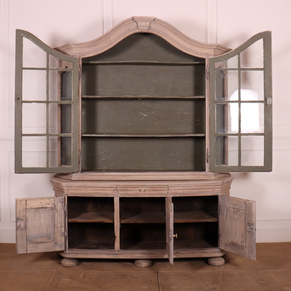 A large Dutch bleached oak display cabinet featuring an arched glazed top section with glass doors and a lower cupboard base supported on bun feet. View VI