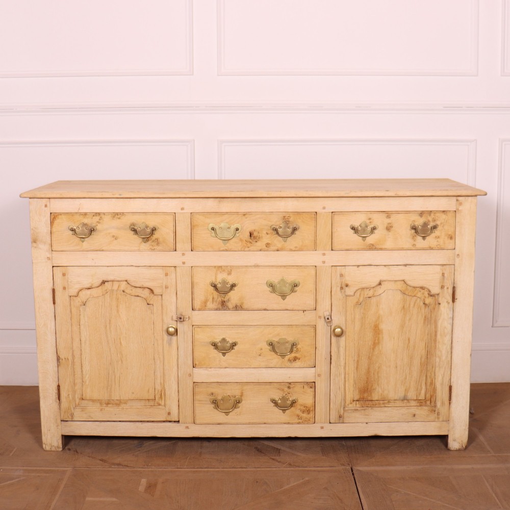 A wide bleached oak dresser base featuring six drawers with brass drop handles and two arched panelled cupboard doors.