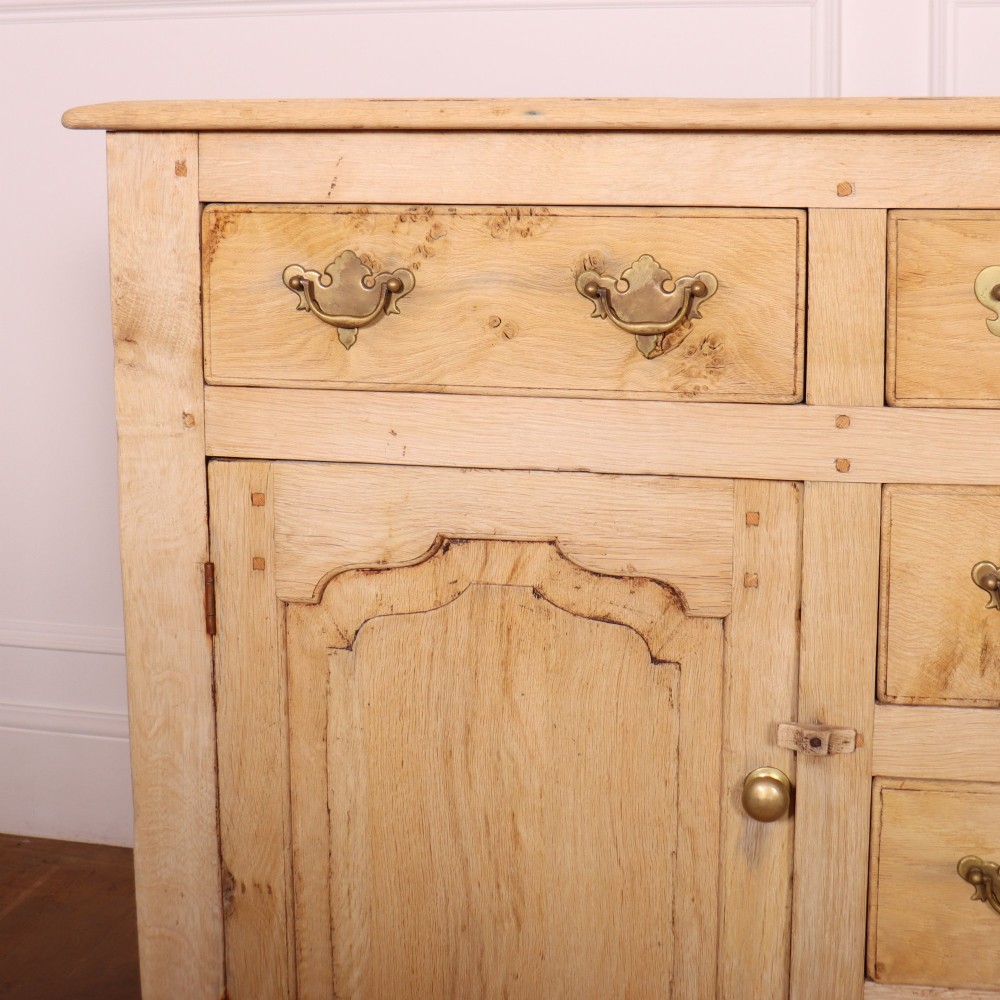 A wide bleached oak dresser base featuring six drawers with brass drop handles and two arched panelled cupboard doors. View II