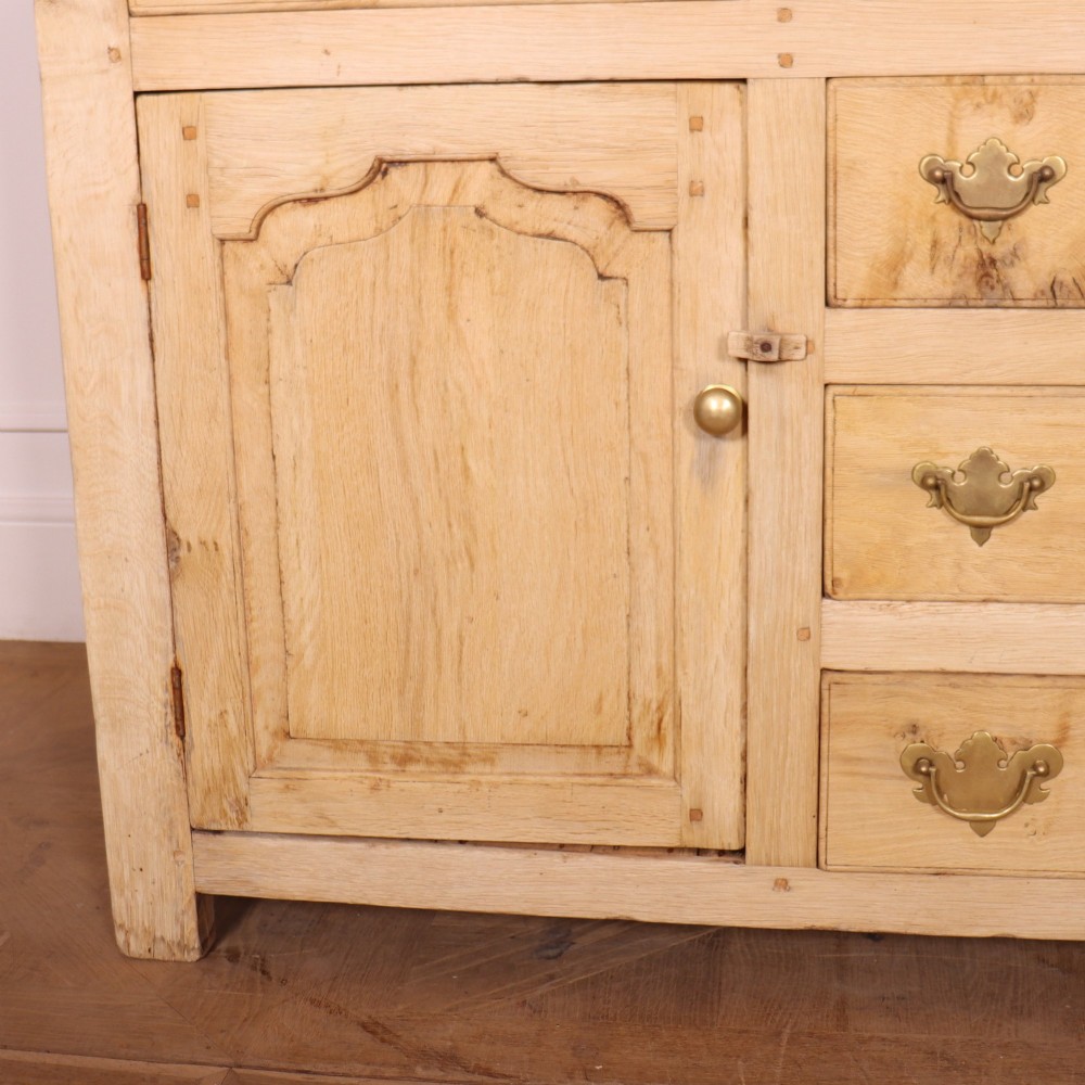 A wide bleached oak dresser base featuring six drawers with brass drop handles and two arched panelled cupboard doors. View III