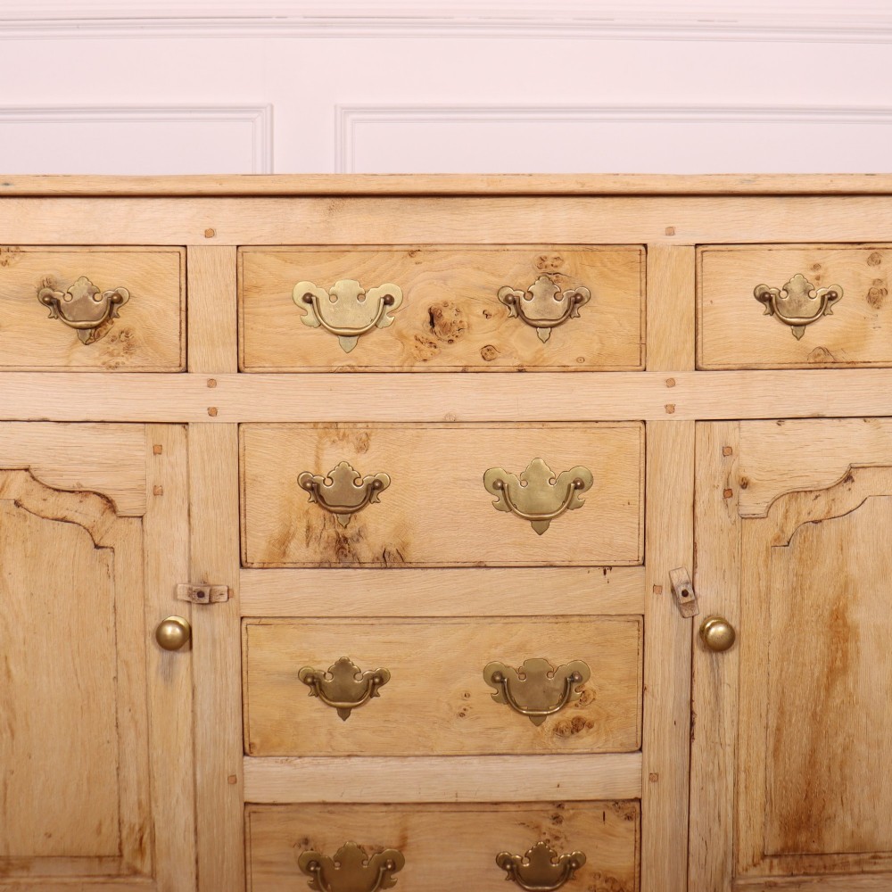 A wide bleached oak dresser base featuring six drawers with brass drop handles and two arched panelled cupboard doors. View IV
