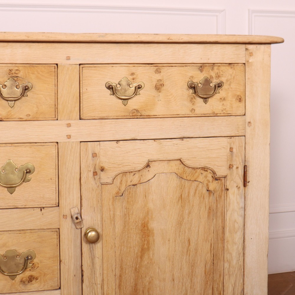 A wide bleached oak dresser base featuring six drawers with brass drop handles and two arched panelled cupboard doors. View V