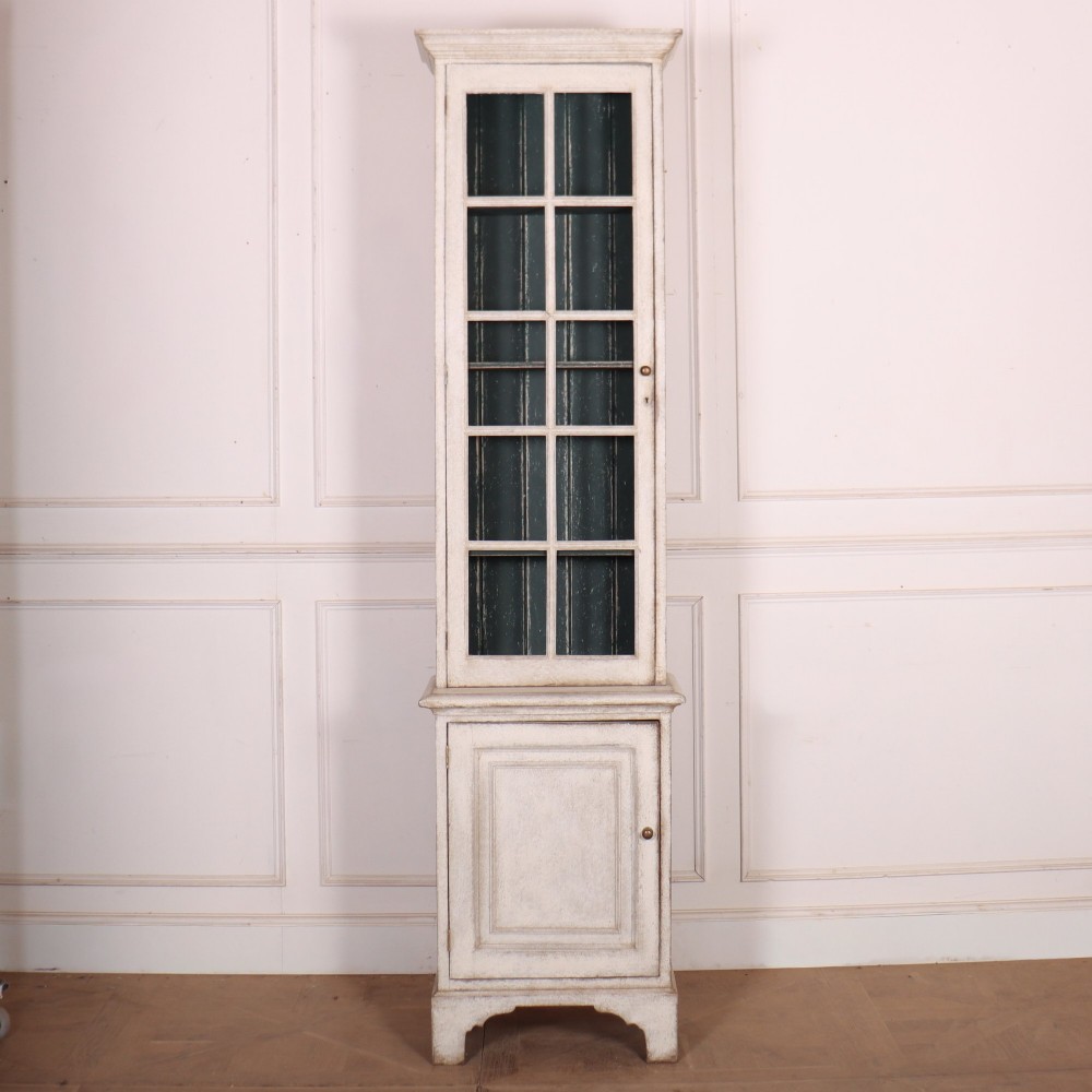 A tall, narrow off-white painted glazed bookcase featuring a glass upper section with a dark interior, above a single panelled cupboard door.