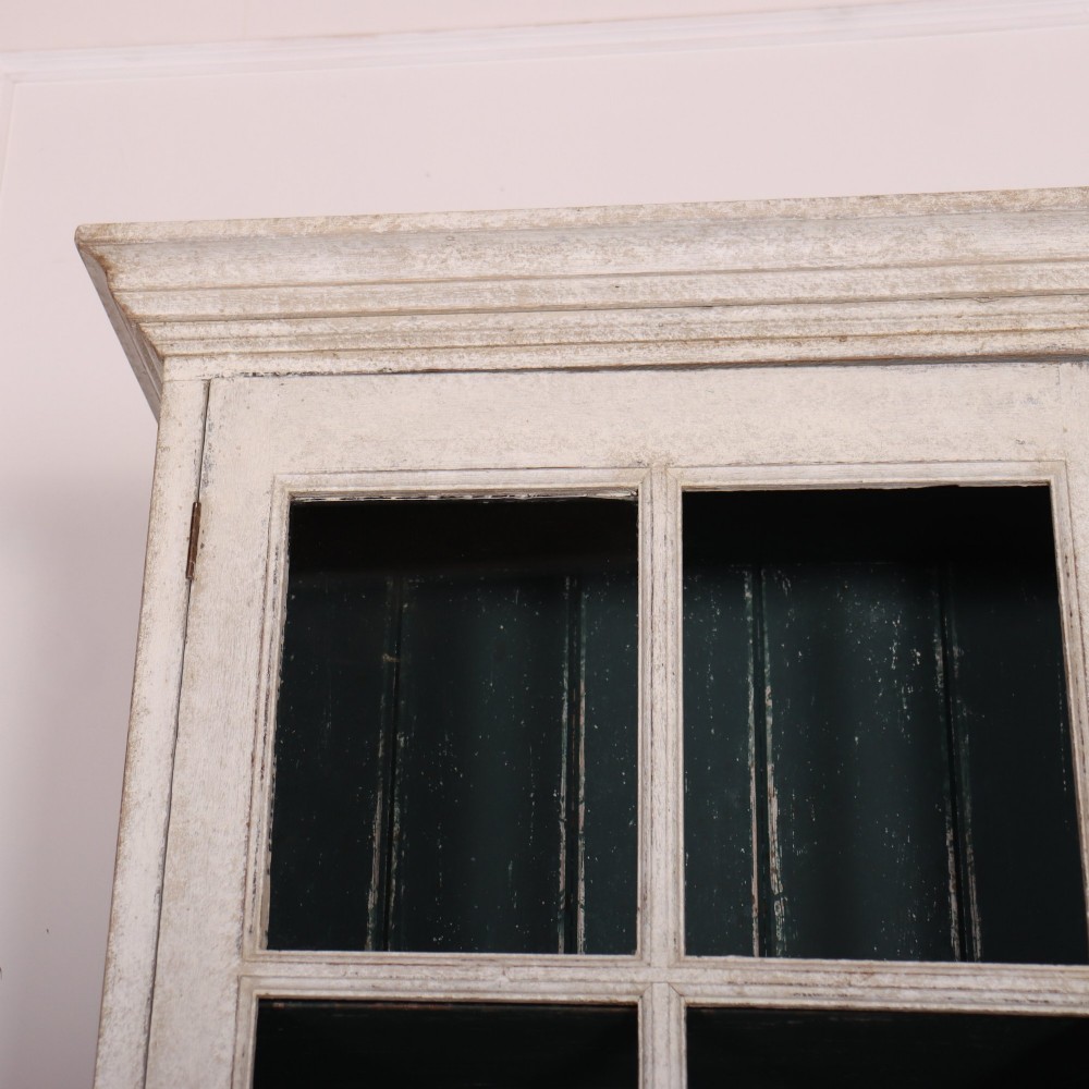 A tall, narrow off-white painted glazed bookcase featuring a glass upper section with a dark interior, above a single panelled cupboard door. View II
