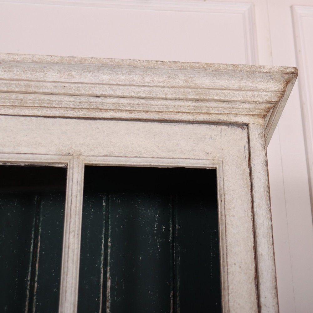 A tall, narrow off-white painted glazed bookcase featuring a glass upper section with a dark interior, above a single panelled cupboard door. View III